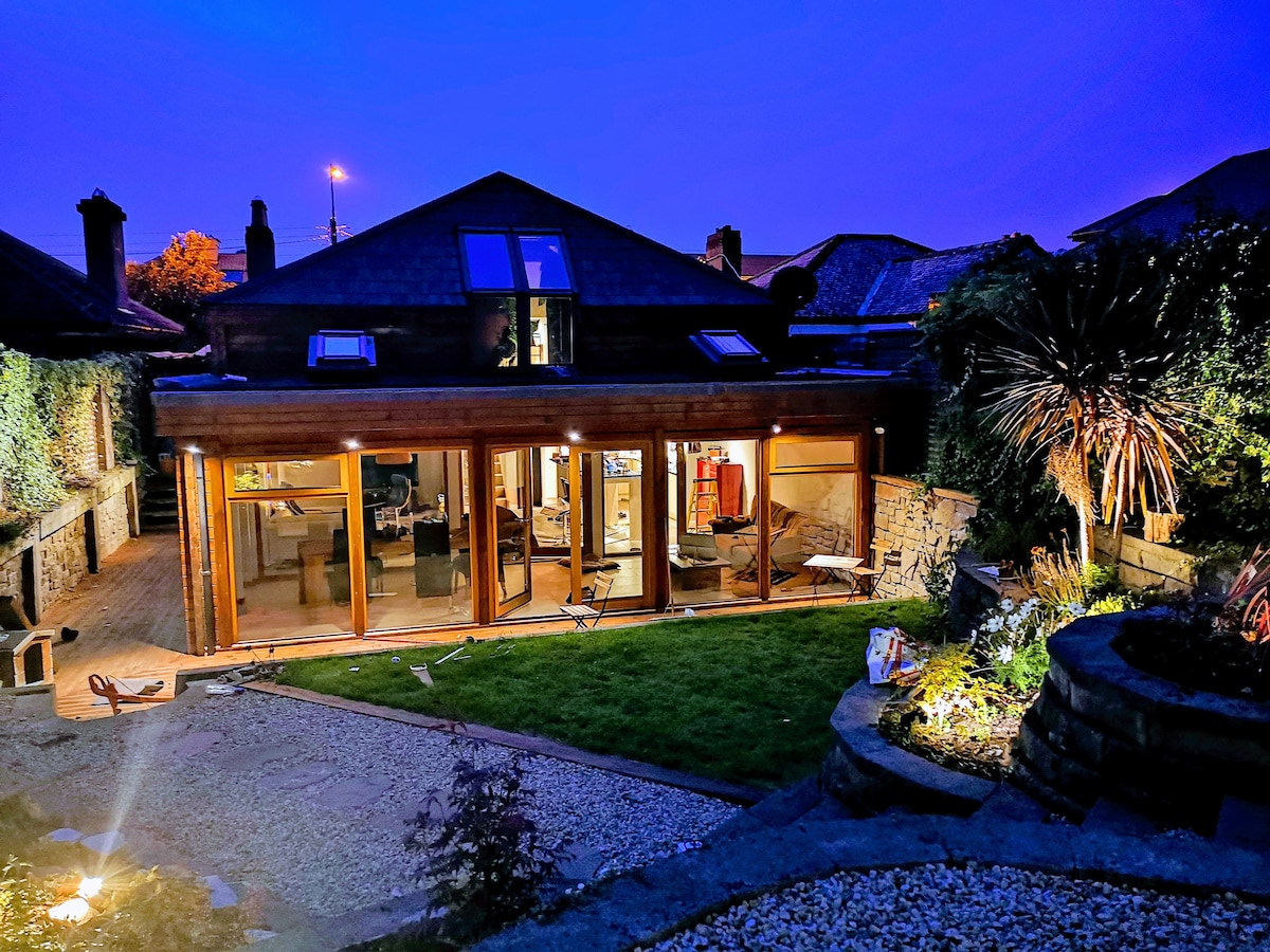 A bungalow with a wooden facade is illuminated at night, showcasing wide glass doors that reveal a spacious interior. The garden features carefully arranged plants and decorative stones, with a pathway leading toward a seating area, providing an inviting outdoor space.