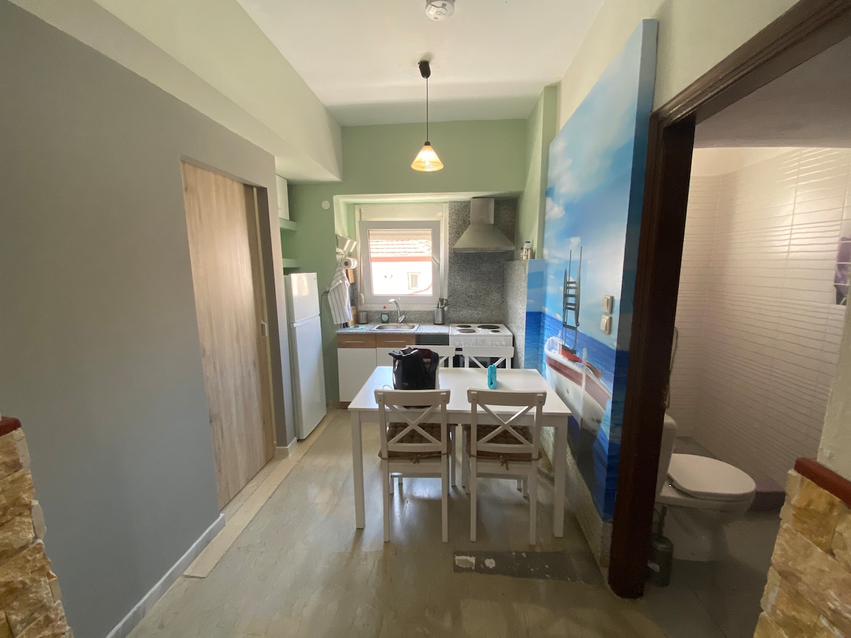 A modern kitchen area features a dining table with white chairs, surrounded by light-colored cabinetry. The kitchen includes essential appliances, with a wall partially adorned by a scenic mural. Natural light enters through a window, creating a bright and functional space.