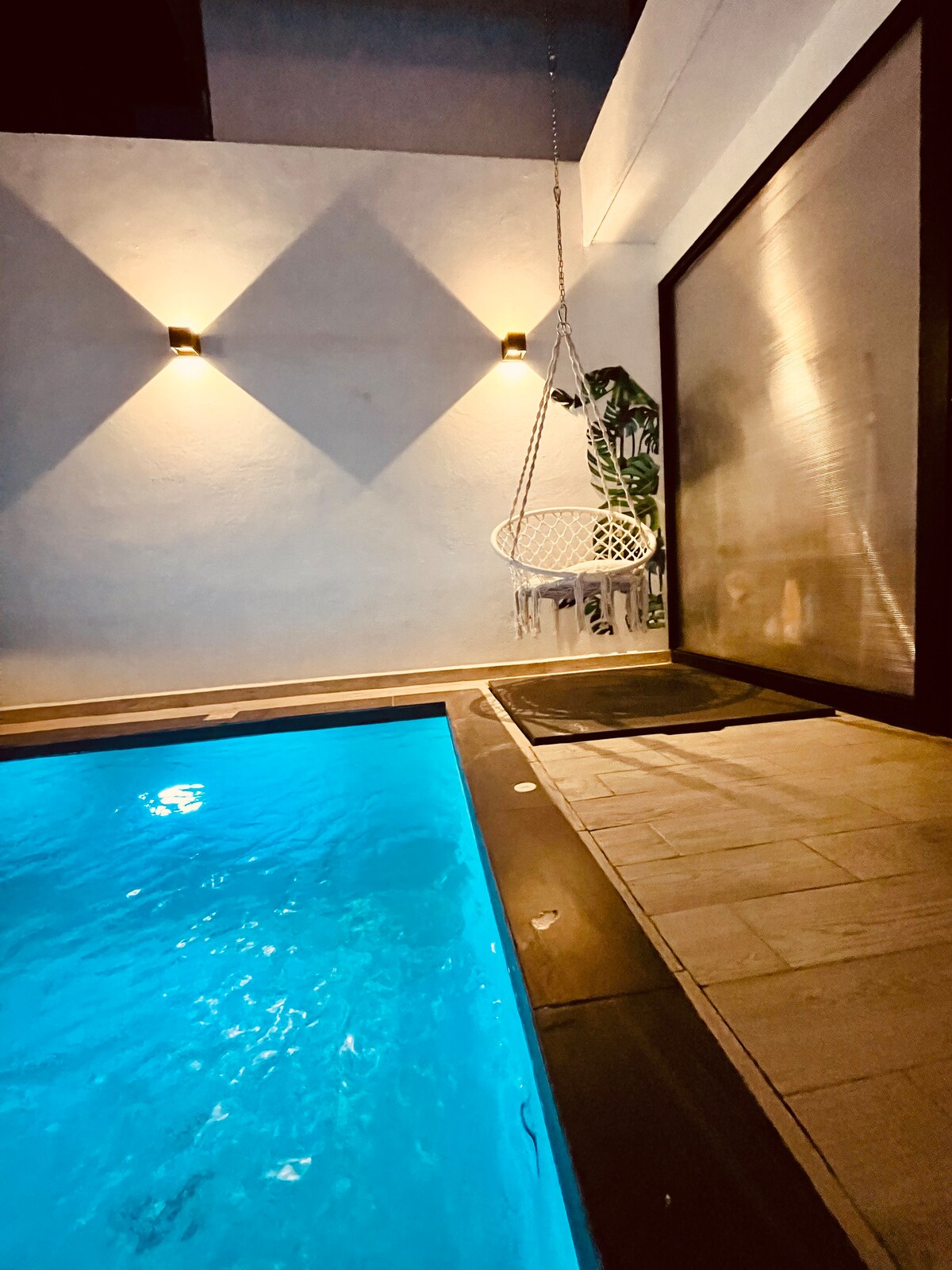 A swimming pool is illuminated with soft blue lighting, reflecting a calm ambiance. A hanging chair is visible beside the pool, providing a relaxation spot. The surrounding walls are minimally decorated, contributing to a serene environment.