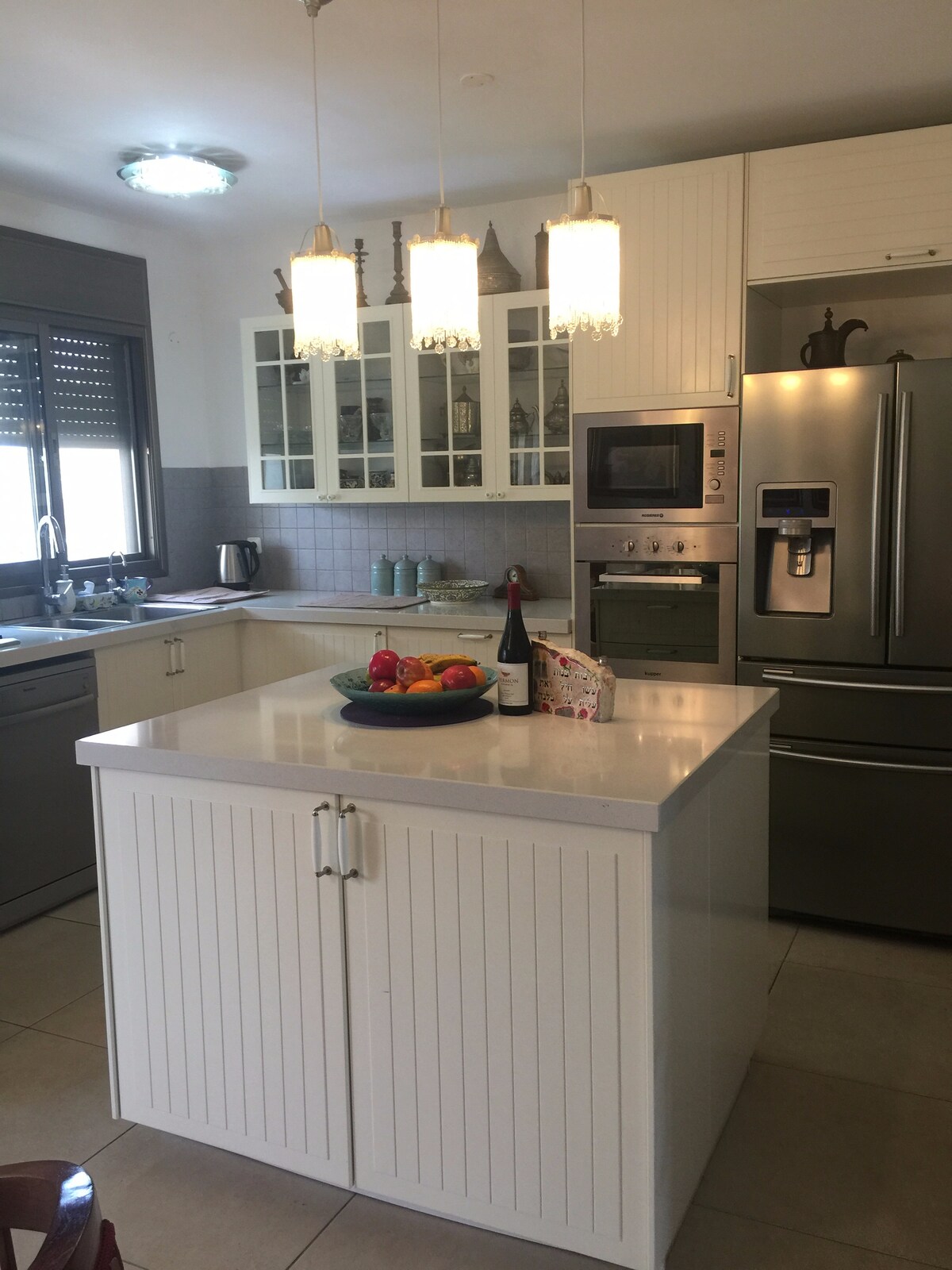 A modern kitchen features a central island with a fruit bowl, illuminated by pendant lights. Cabinets display glassware, and stainless steel appliances are arranged, including a microwave and refrigerator. Natural light enters through a window adorned with horizontal blinds, enhancing the airy feel.