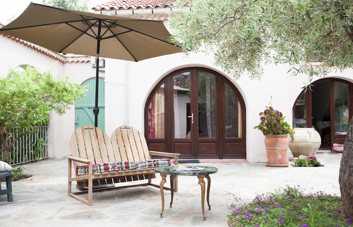 An outdoor seating area features two wooden lounge chairs with cushions alongside a small round table. A large umbrella provides shade, complemented by greenery and potted plants. The background includes arched doorways leading into the house.