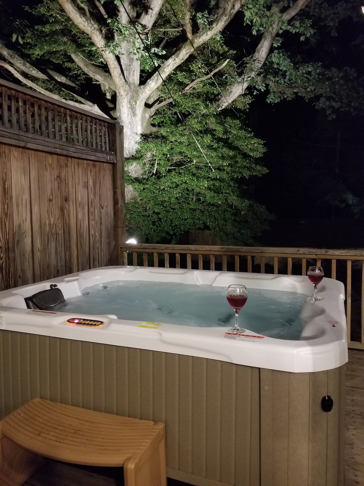 A hot tub is positioned on a wooden deck, surrounded by tall trees and softly lit fixtures, creating a serene ambiance. Two glasses of red liquid rest on the edge of the tub, inviting relaxation under the night sky.