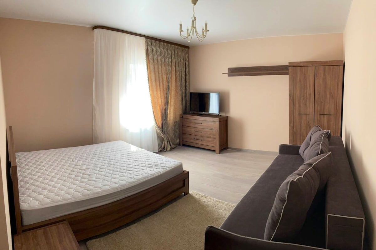 A spacious room features light peach walls and natural light streaming through a large window. A bed frame stands ready for a mattress, accompanied by a brown sofa set. A dresser and TV on a cabinet add functional elements to the inviting space.