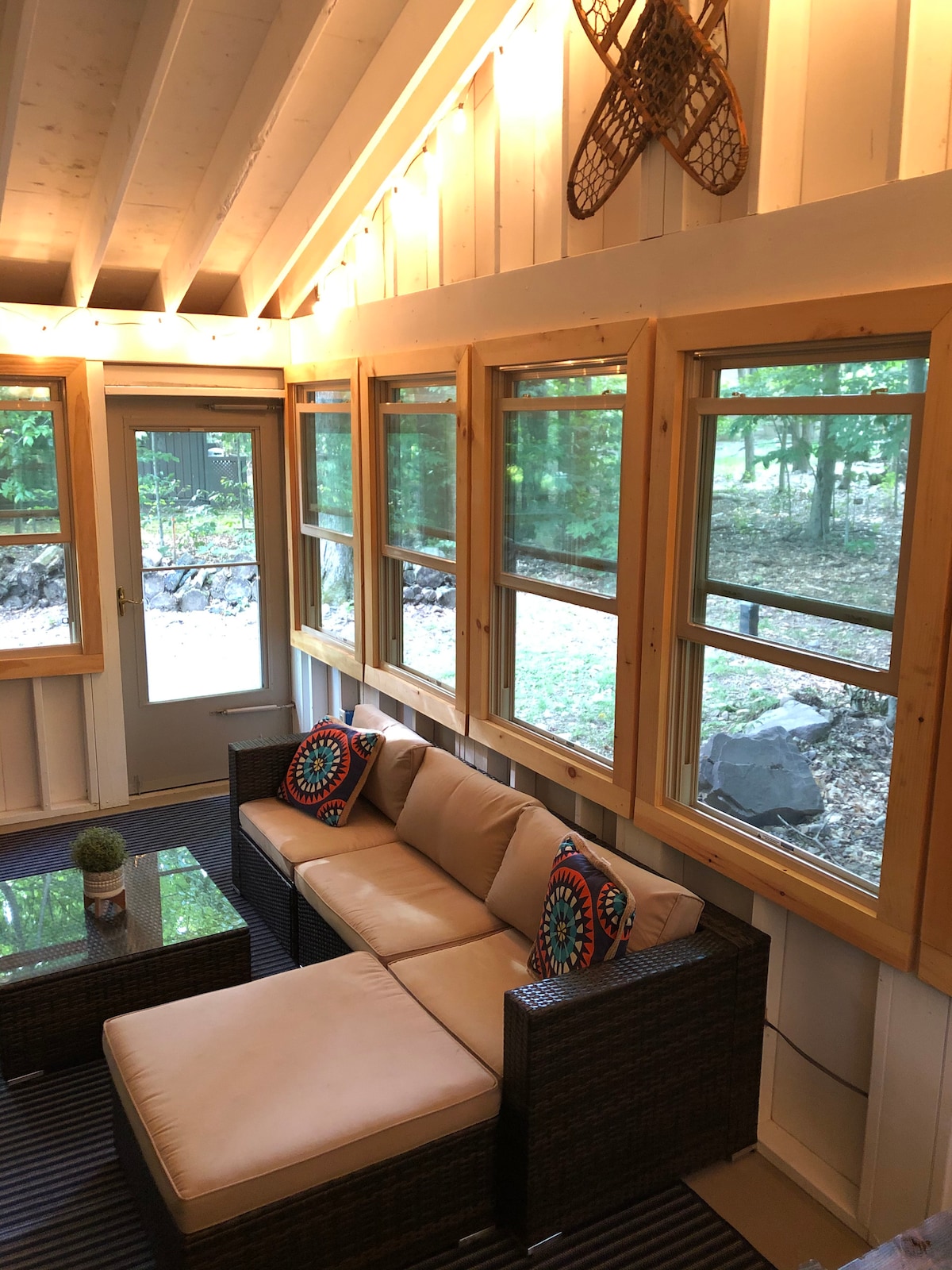 A sunroom is showcased with large windows allowing natural light to enter. A comfortable seating area features a light-colored sofa with decorative cushions, complemented by a low table. The walls are adorned with wooden accents, enhancing the cozy ambiance.