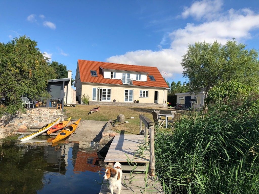 A modern house is situated by the shore, with a red-tiled roof and multiple large windows reflecting the sky. Kayaks are neatly arranged along the water's edge, and a path leads to a dock. Lush greenery frames the scene, enhancing the natural environment.