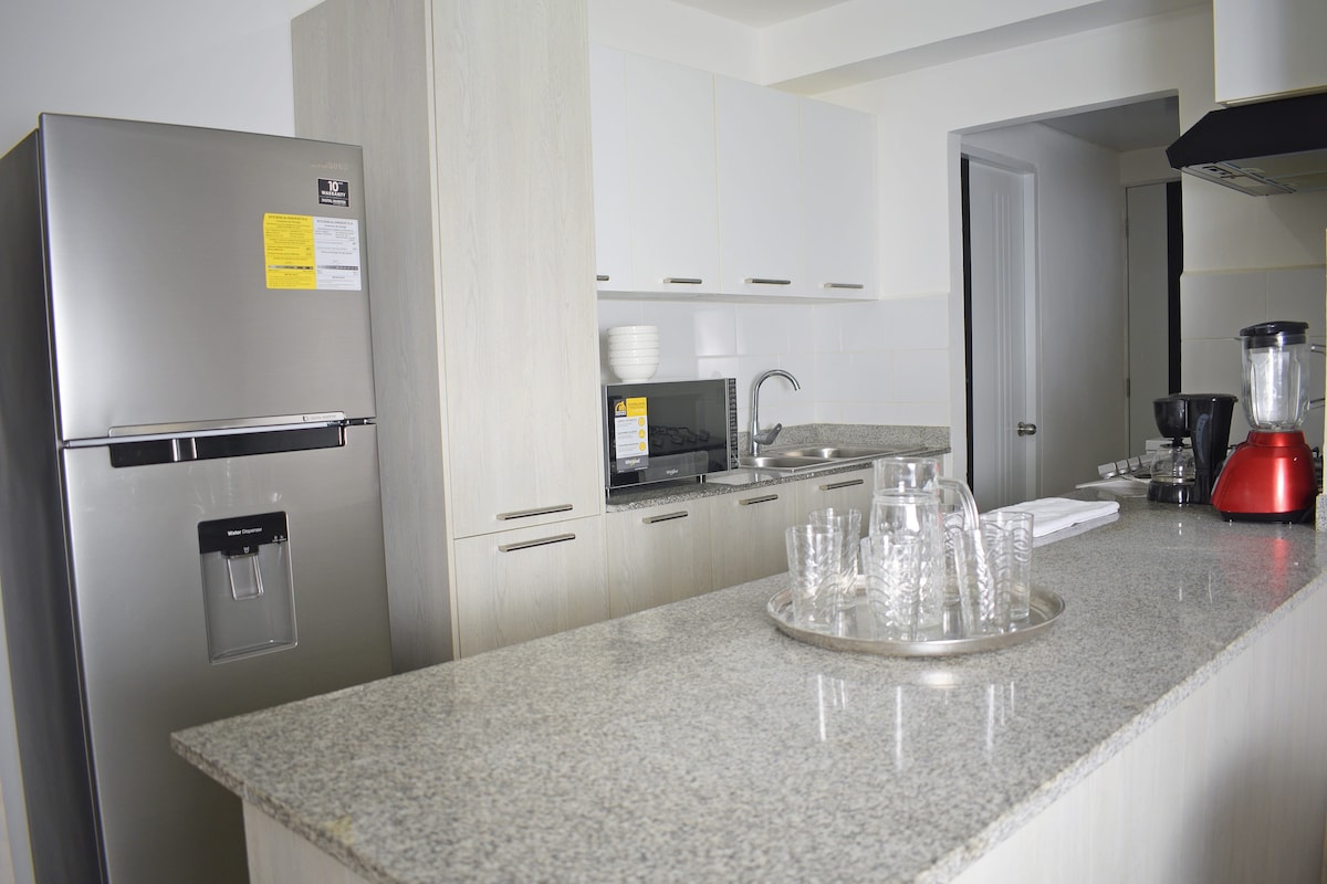 A modern kitchen is equipped with stainless steel appliances, including a refrigerator and blender. Counter space is available with a textured granite surface. Glassware is neatly arranged on the counter, and cabinets above provide additional storage. The kitchen opens to a bright area with natural light.