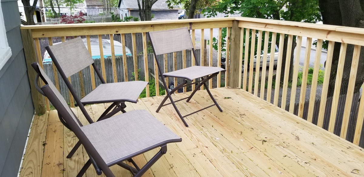 A private wooden patio features three foldable chairs arranged for relaxed seating. The space is surrounded by a wooden railing, offering views of greenery beyond. The weathered wooden deck shows signs of nature with scattered leaves.