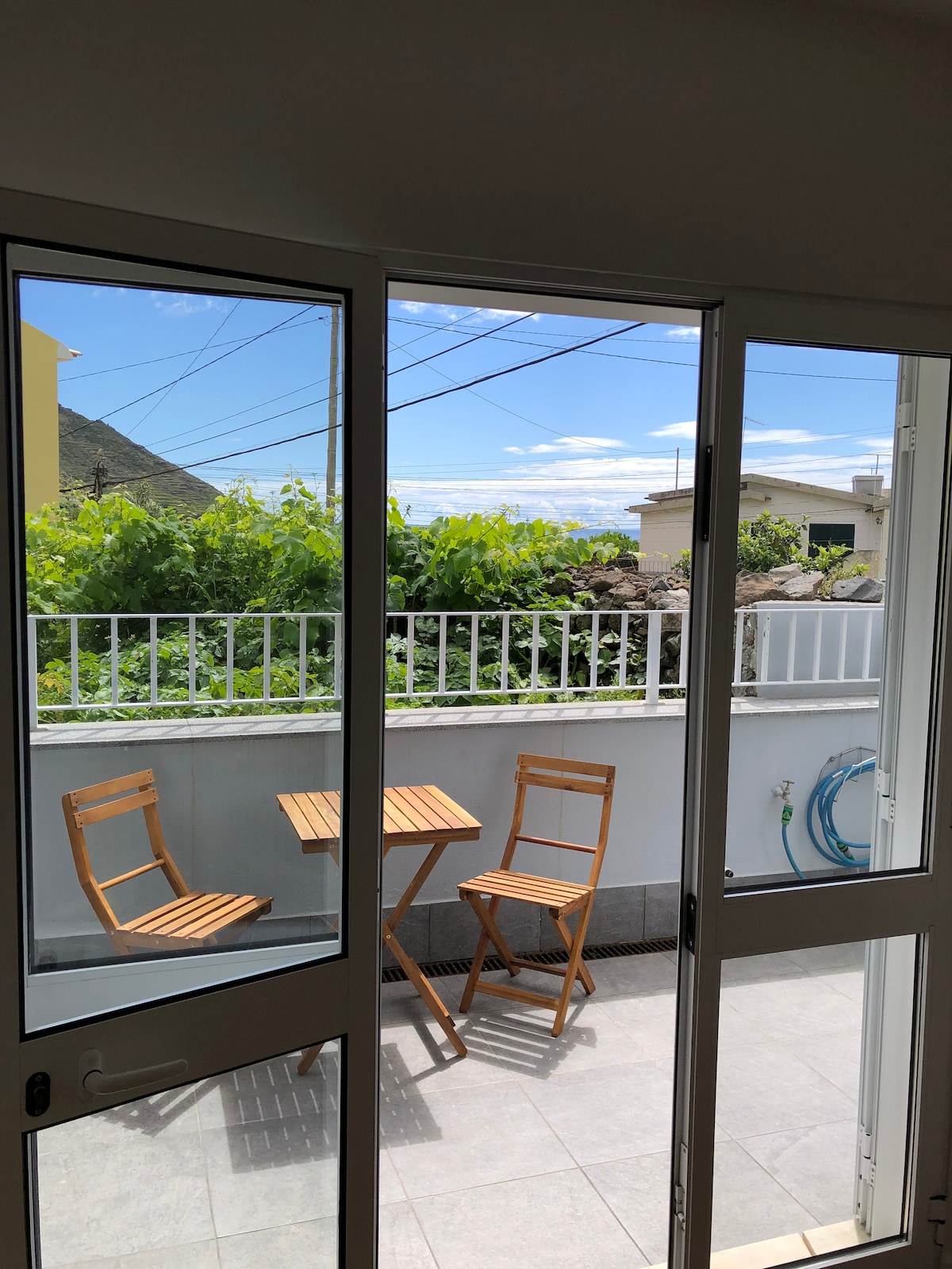 A view from the open patio doors reveals a small outdoor area with a table and two folding chairs. Lush greenery and a blue sky create a refreshing backdrop, while mountains are visible in the distance.