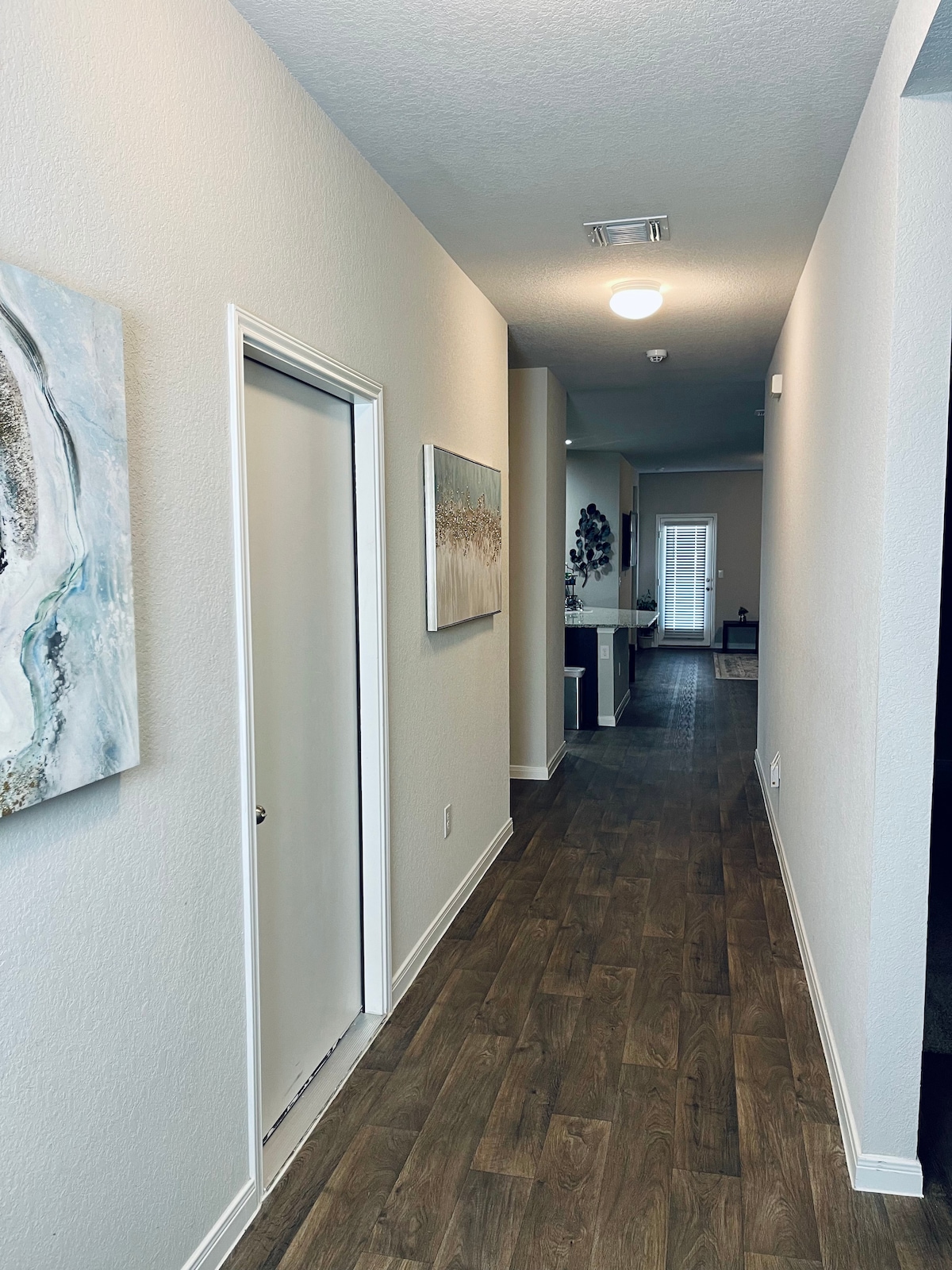 A well-lit hallway is shown, featuring smooth, wooden flooring and neutral-colored walls. Artwork decorates the left side, while a door is visible on the left. At the end of the hall, a glimpse of light suggests an open area or room beyond.