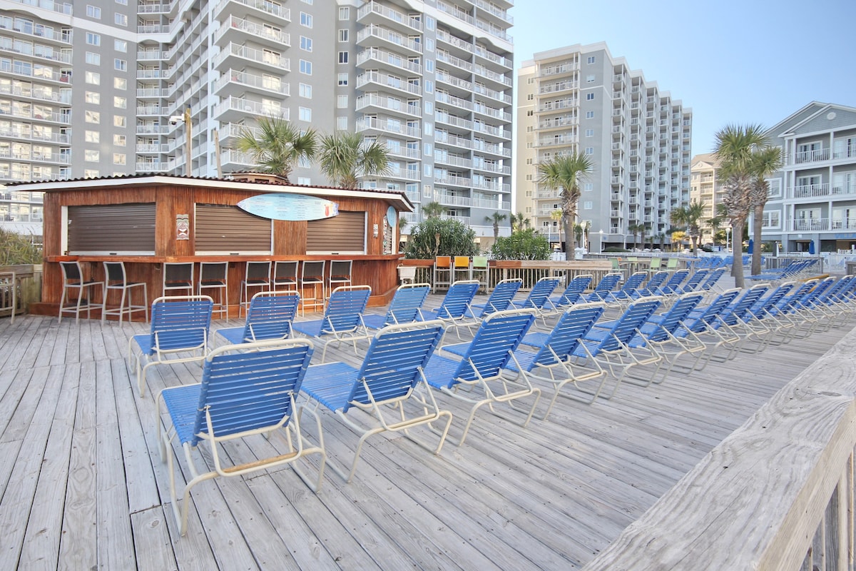 A spacious deck area features numerous blue lounge chairs arranged in neat rows, facing a Tiki bar structure made of wood. The backdrop includes tall residential buildings and palm trees, complemented by a clear sky.