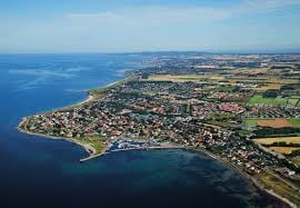 An aerial view of a coastal area shows a variety of landscapes, including residential neighborhoods, agricultural fields, and the shimmering blue sea. The coastline curves gently, and several boats are visible in the harbor, providing a glimpse of the vibrant maritime activity.