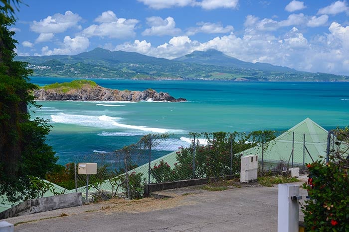 A scenic coastal view showcases the turquoise sea and distant mountains under a partly cloudy sky. The foreground features a paved path framed by greenery and shrubs, leading towards the vibrant water and rugged shoreline, creating an inviting atmosphere for exploration.