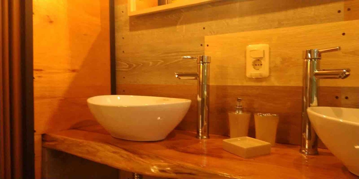 A modern bathroom sink area is displayed with a white bowl sink resting on a smooth wooden countertop. Sleek chrome faucets are positioned beside two glass containers and a small, square soap dish, all set against a textured wooden wall.