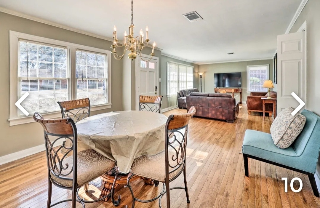 A spacious living area features a circular dining table surrounded by elegant chairs. Natural light pours through large windows, illuminating the wood flooring. A comfortable seating arrangement is visible in the background, complemented by a flat-screen TV and a decorative lamp.