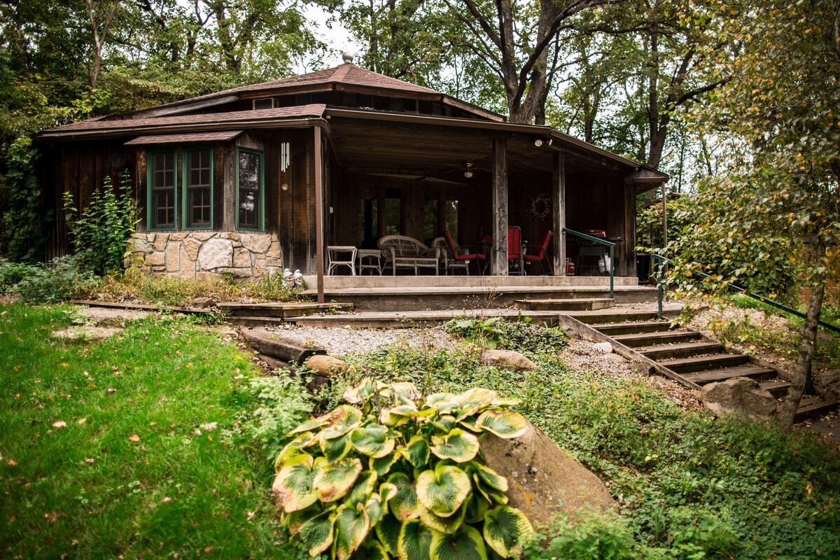 The rustic lodge, surrounded by greenery, features a spacious porch with seating options. Steps lead up to the entrance from the landscaped yard, which includes various plants and rocky elements. Natural wood tones are visible in the structure, enhancing the outdoor integration.