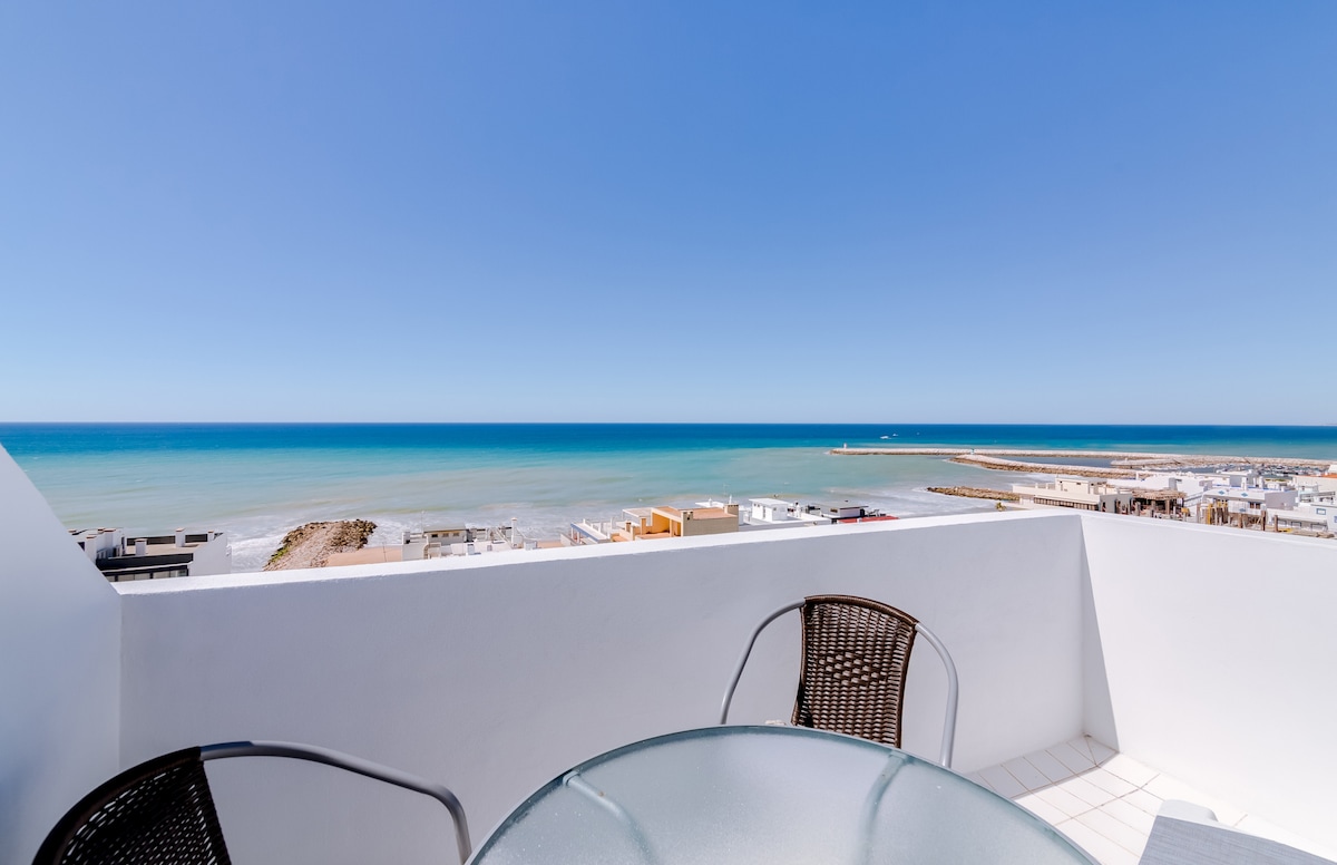 A terrace view showcases the expansive sea, with calm waters meeting the horizon. Sunlight reflects gently on the surface, while two wicker chairs are positioned around a glass table, creating a relaxed outdoor sitting area for enjoying the coastal scenery.