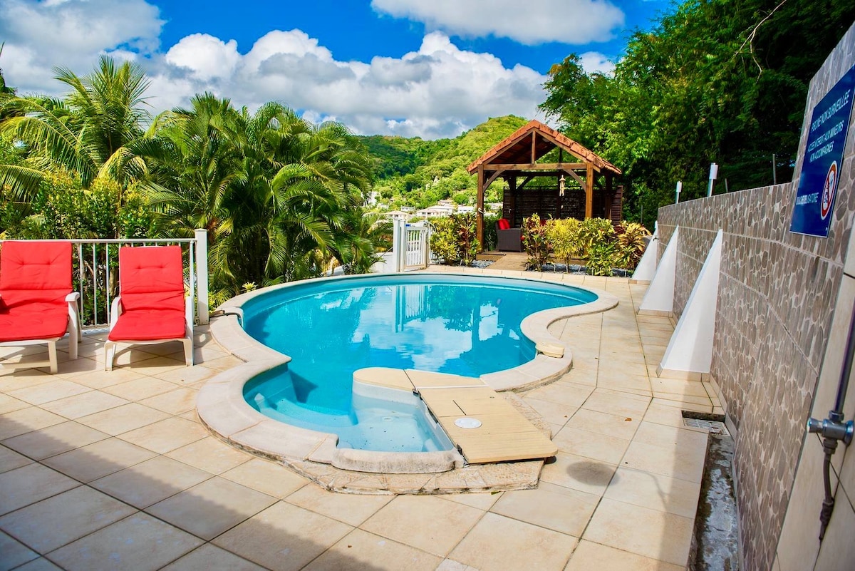 A private pool area features a uniquely shaped heated pool surrounded by a tiled deck. Two red lounge chairs are positioned near the pool, and a gazebo is visible in the background, nestled among tropical greenery. The scene is complemented by a partly cloudy sky.