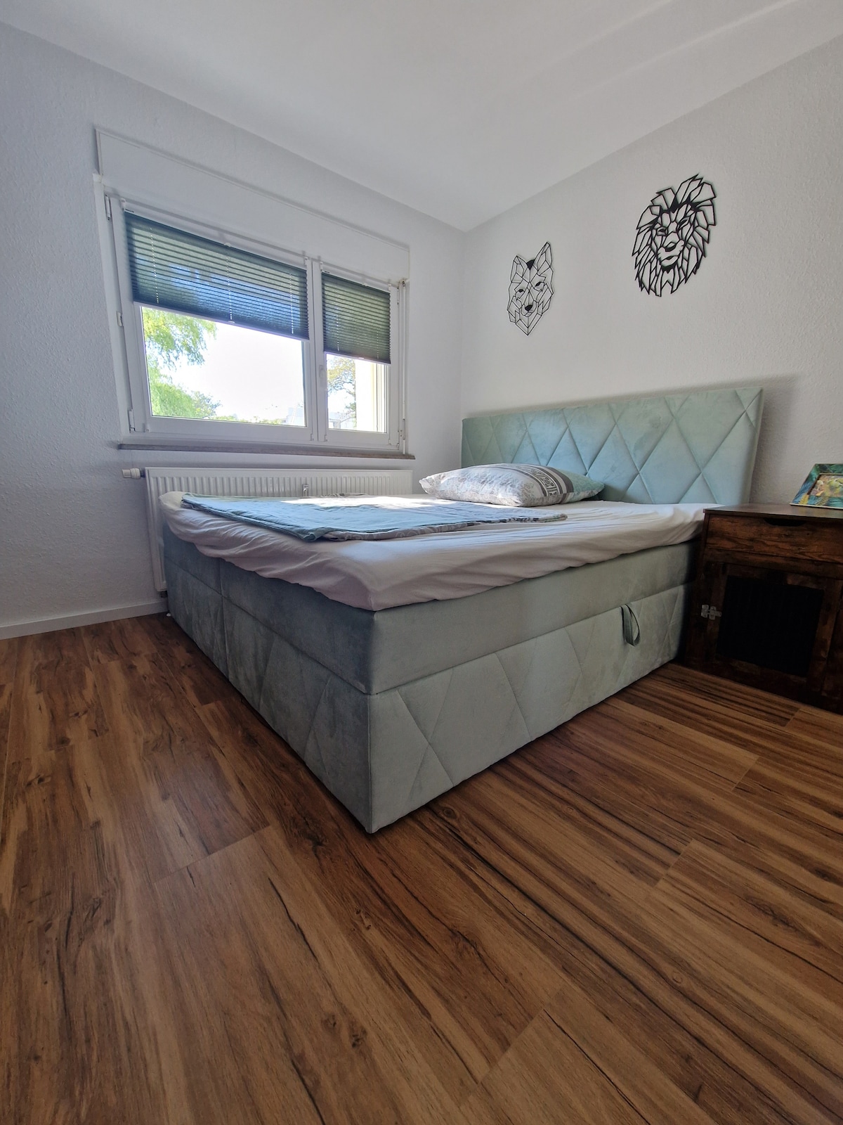The bedroom features a spacious bed with a soft, upholstered headboard and a neatly made bedspread. Natural light enters through two windows, adding a fresh, airy feel. The wooden floor complements the calming decor, highlighted by decorative animal-themed wall art.
