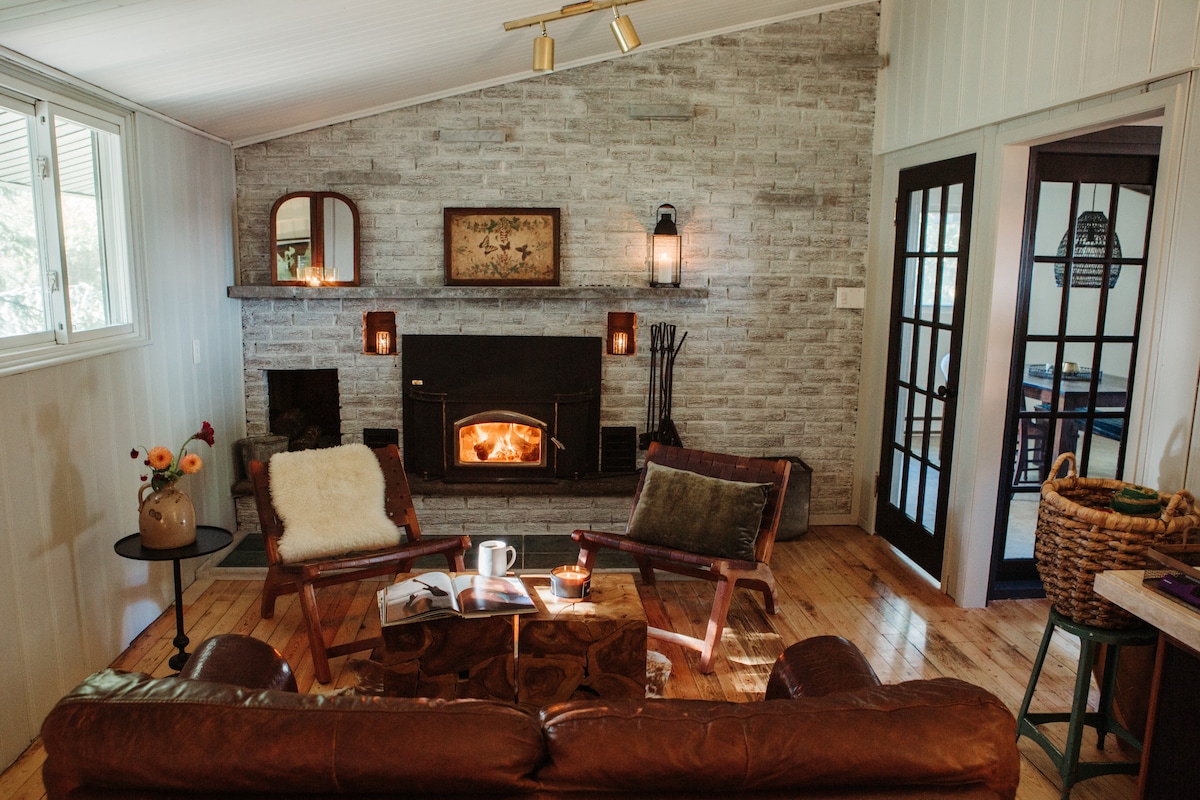 A warm living area features a stone fireplace as the focal point, with a cozy fire burning inside. Comfortable leather seating is arranged around a rustic wooden coffee table, complemented by small decorative items and soft textiles, while glass doors lead to another space.