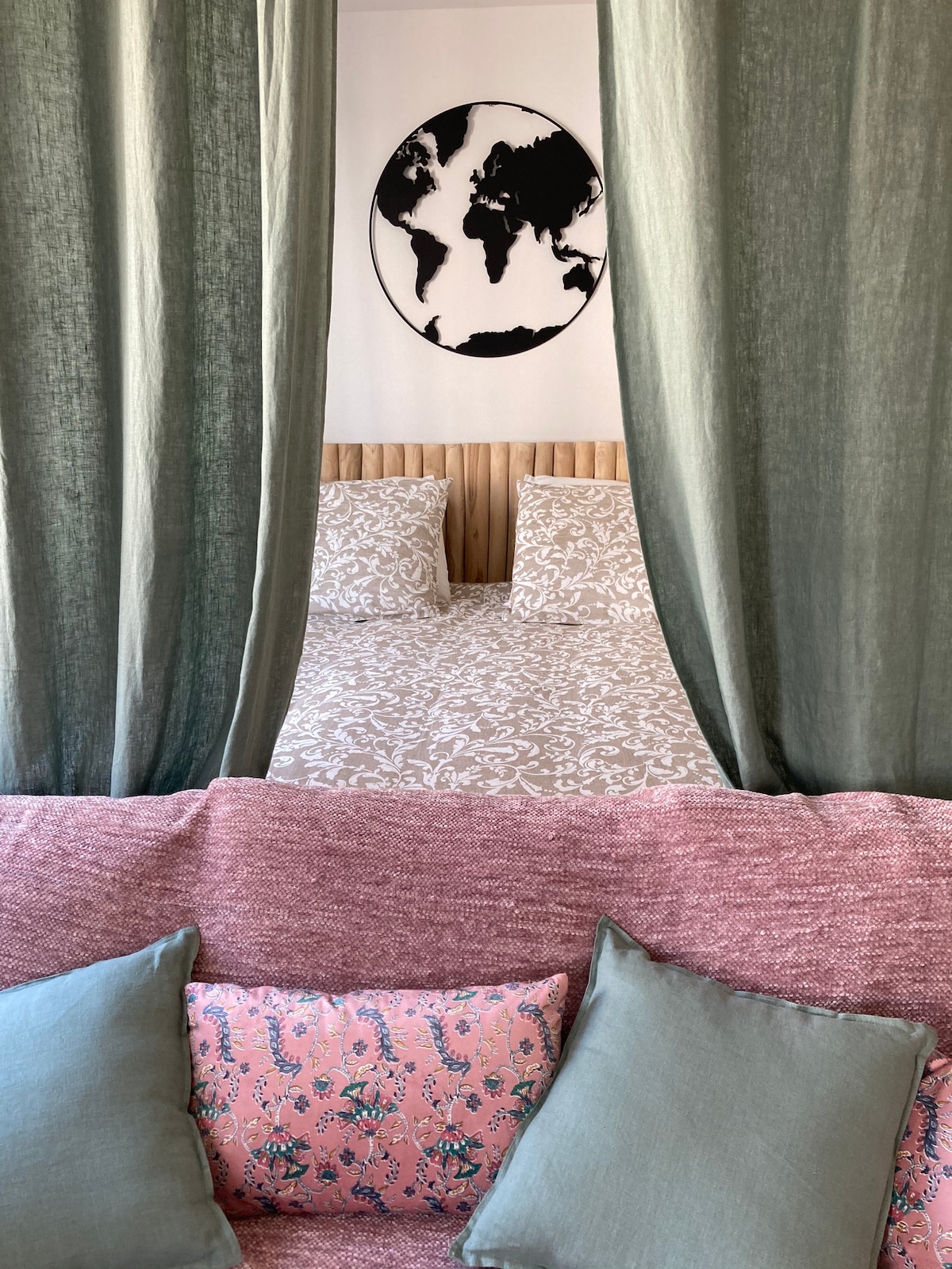 A cozy sleeping area is defined by flowing green curtains that frame a spacious bed covered with a patterned duvet. A decorative wall art piece featuring a world map adds interest above the bed. A pink couch with patterned cushions offers additional seating.