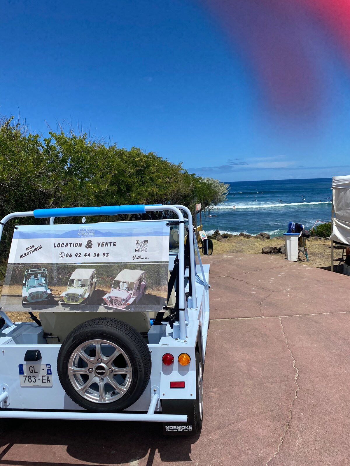 A light blue vehicle is parked on a pathway with a view of the ocean. The back of the vehicle displays a sign advertising rentals, featuring images of colorful cars. Lush greenery and a sandy beach are visible in the background.