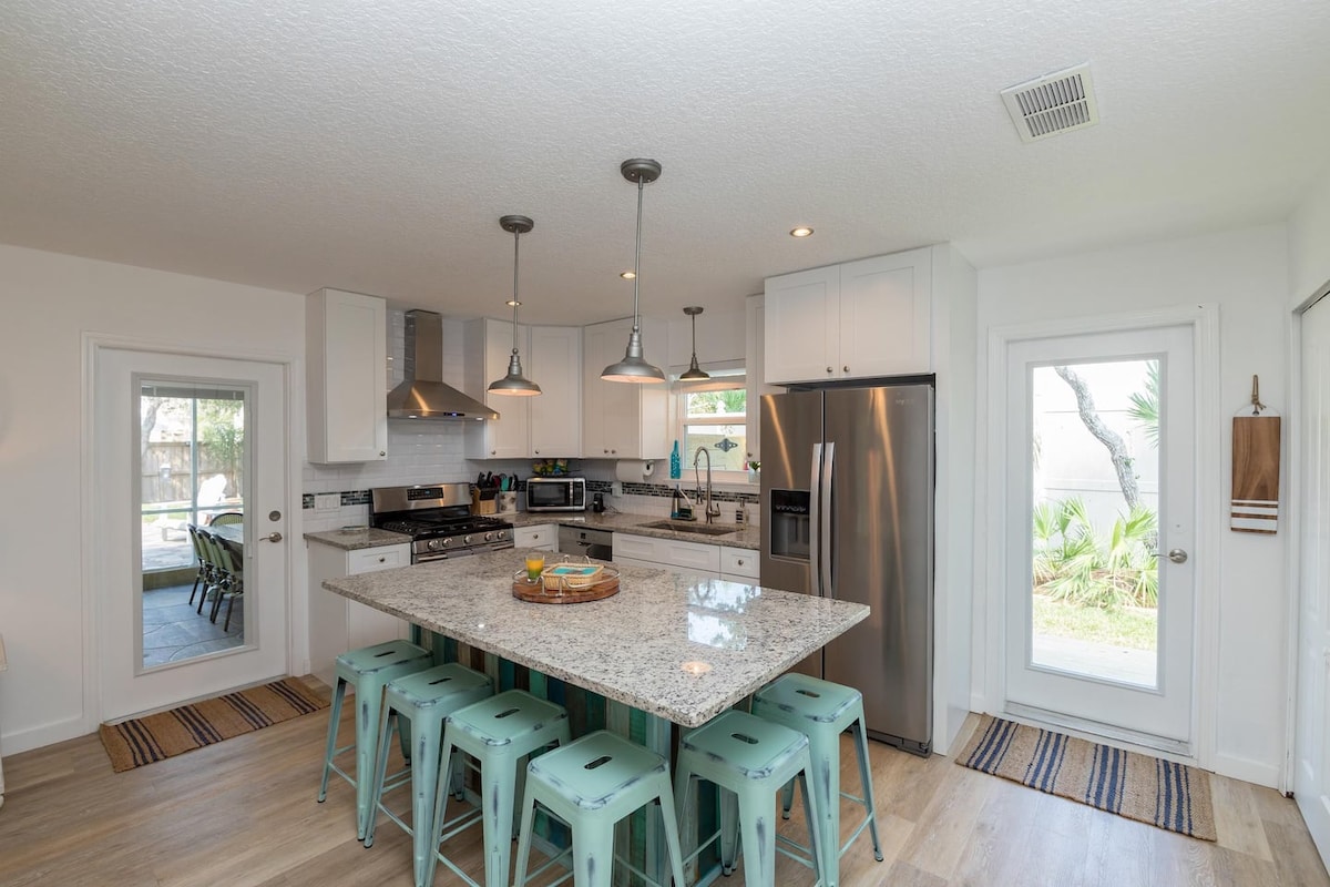 The kitchen features bright white cabinetry and modern stainless steel appliances. A large, central island with a granite countertop is surrounded by turquoise stools. Natural light floods the space through a sliding glass door leading to the outdoor area, creating an inviting atmosphere.