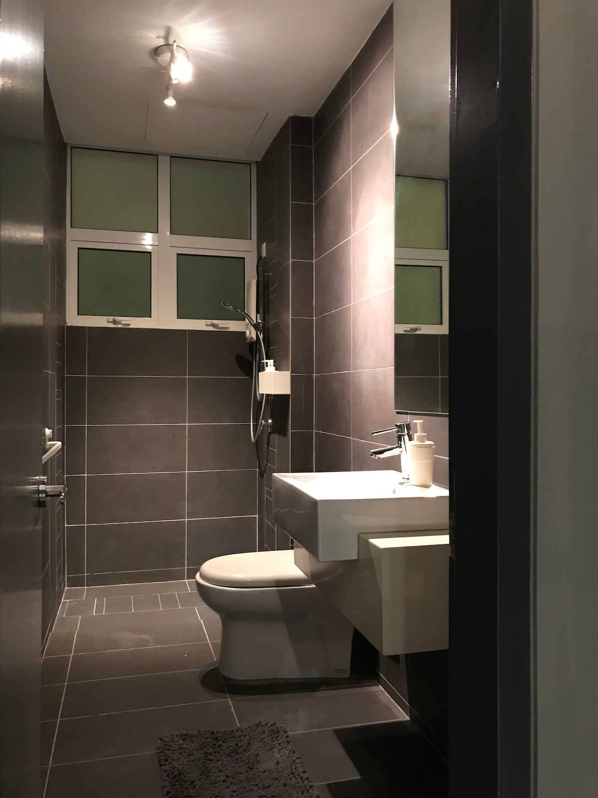 The bathroom features contemporary design elements with dark gray tile walls. A modern sink and toilet are positioned against the wall. Natural light enters through two small windows, enhancing the space's brightness. A textured rug is placed in front of the sink area.
