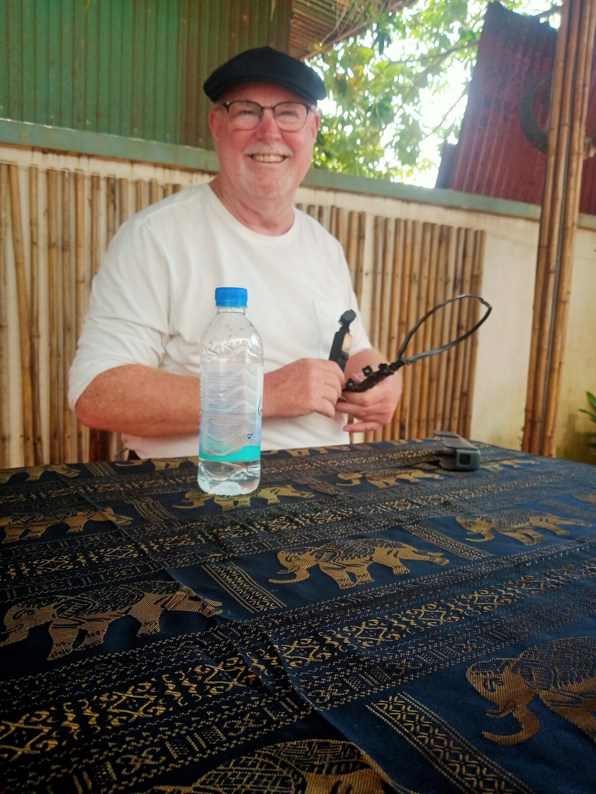A table draped with an intricately patterned fabric featuring elephants sits in a shaded area. A clear water bottle is positioned in front, while a person is seated, holding a pair of glasses. Natural greenery is visible in the background, creating a serene environment.