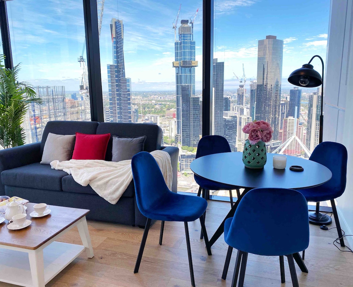 The living area features a comfortable gray sofa adorned with a beige throw and red accent pillow. A dining table is surrounded by four blue chairs, with a decorative plant beside it. Large windows provide expansive city views, flooding the space with natural light.
