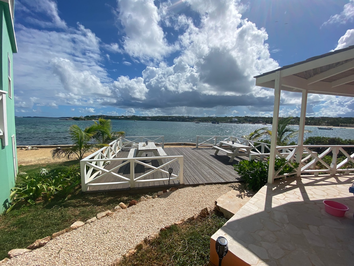 A private deck stretches over the water, framed by tropical greenery. Outdoor seating with white tables and chairs invites relaxation while offering unobstructed views of the sea. The scene features a mix of rocky and sandy beach areas under a partly cloudy sky.