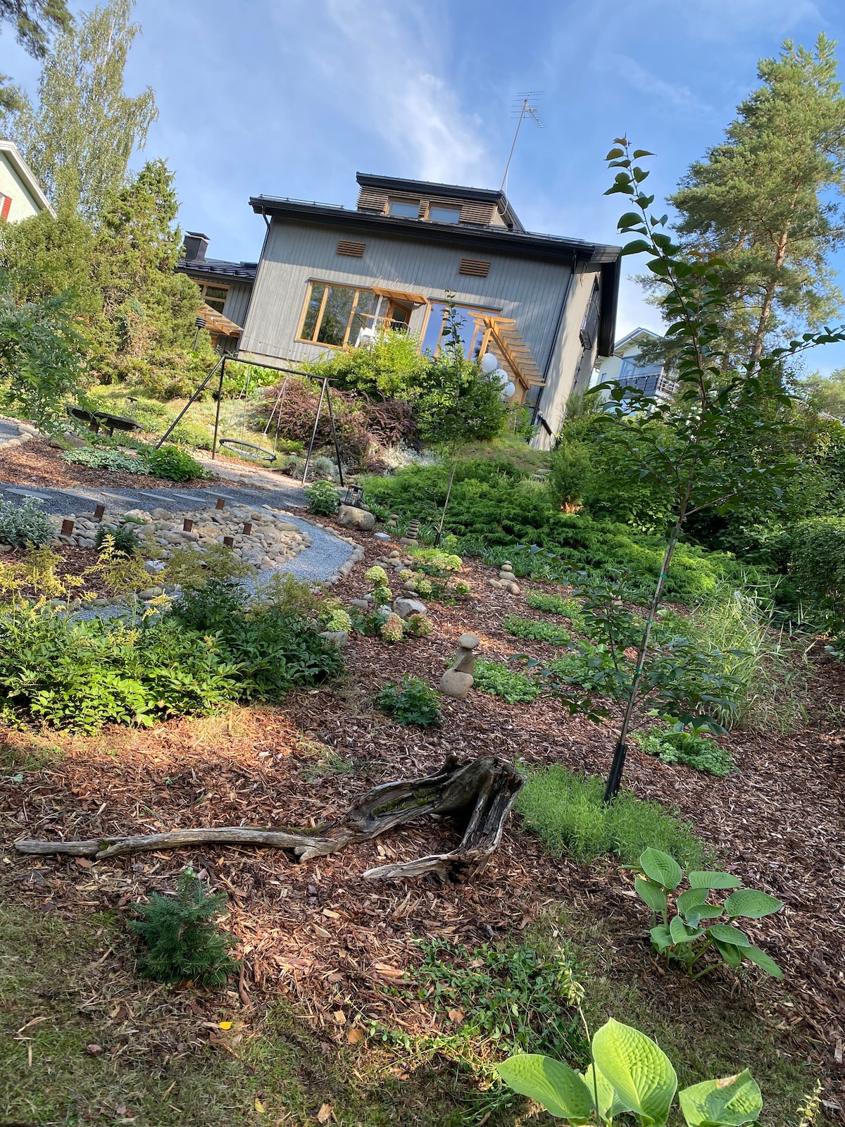 A multi-story house is visible amidst a lush garden, with natural stone pathways and various plants lining the hillside. The arrangement of greenery includes shrubs and trees, creating a sense of tranquility in the outdoor space under a clear blue sky.