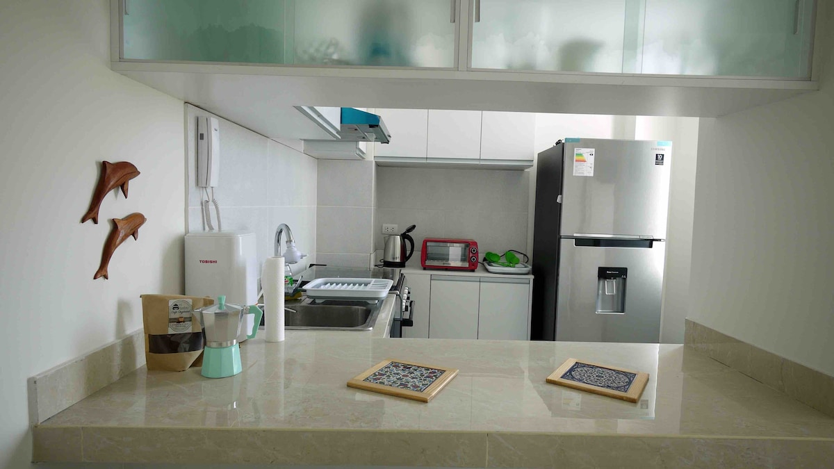 A compact kitchen area is presented with modern appliances, including a refrigerator and a microwave. A sink with a stove is positioned against a white cabinetry backdrop. Two placemats are placed on the countertop, with an espresso maker and kitchen utensils visible nearby.