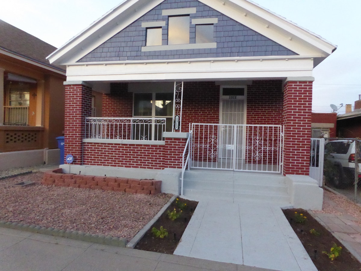 The exterior of a bungalow-style house is displayed, featuring a brick façade and a front porch with decorative railings. A concrete walkway leads to the entrance, bordered by low landscaping. The home is situated in a quiet neighborhood, reflecting a welcoming approach.