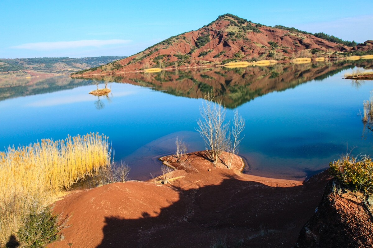 A calm lake is surrounded by hills with reddish-brown terrain. Lush grass and yellow reeds line the shore, reflecting in the still water. Gentle slopes lead to the water's edge, creating a serene and tranquil atmosphere in the natural landscape.