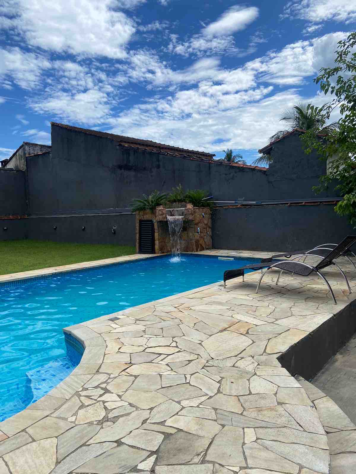 An inviting swimming pool is shown, framed by a stone patio. A water feature cascades gently into the pool, while lounge chairs are positioned along the edge. Lush green grass enhances the outdoor space, complemented by a backdrop of tall trees and a blue sky.