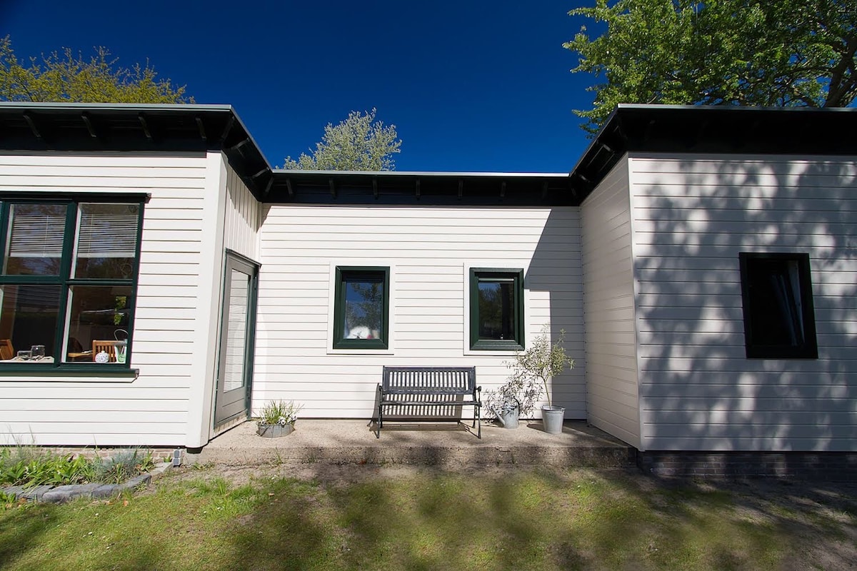 The exterior of a modern, single-story house is shown, featuring a clean white facade and large windows. A wooden bench is positioned on a small patio area, providing a space for outdoor relaxation. Lush greenery surrounds the house, creating a serene environment.