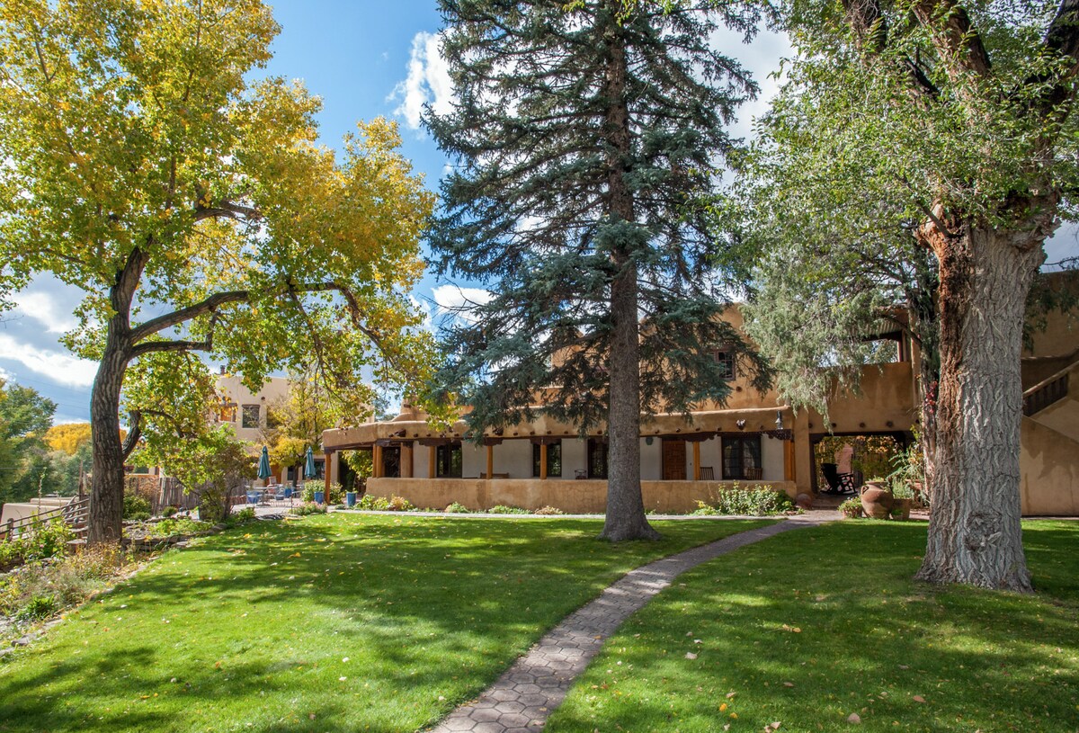A lush garden area is framed by mature trees, featuring a winding stone pathway that leads past the adobe-style building. The grounds are manicured, showcasing a vibrant mix of greenery and seasonal foliage under a bright blue sky with scattered clouds.