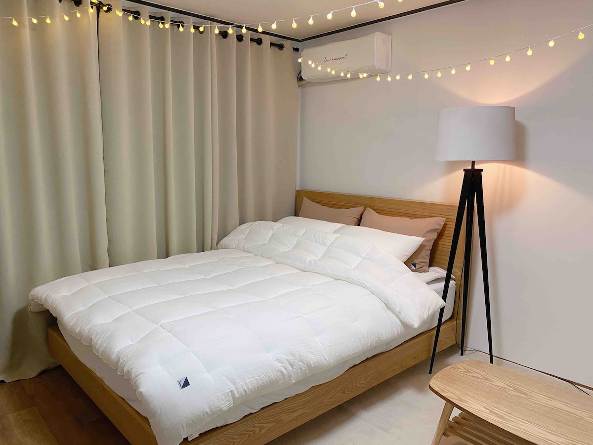 A spacious bed with a soft comforter is positioned against a neutral wall. Soft curtains frame the window, while delicate string lights add subtle illumination. A floor lamp stands beside the bed, contributing to the calming ambiance of the room.