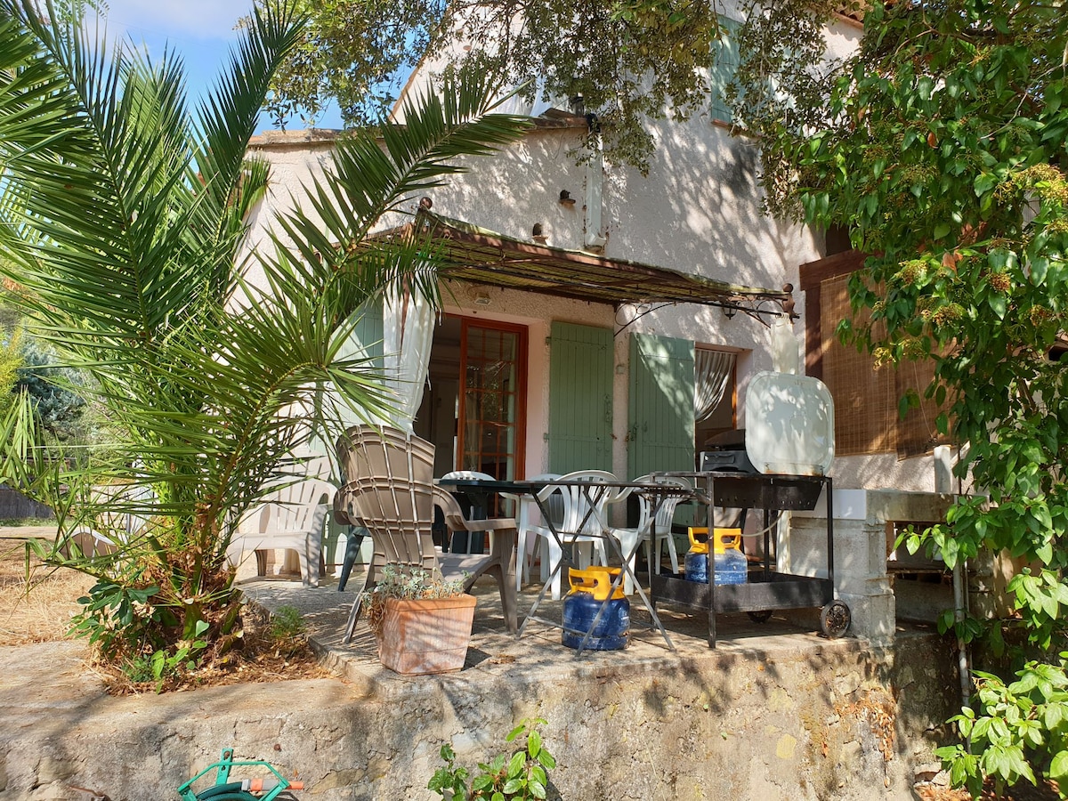 A terrace area is surrounded by lush greenery and features a dining table with chairs. A barbecue station is visible, and shaded seating arrangements invite relaxation. The exterior of the building is light-colored, with traditional shutters and a stone foundation.