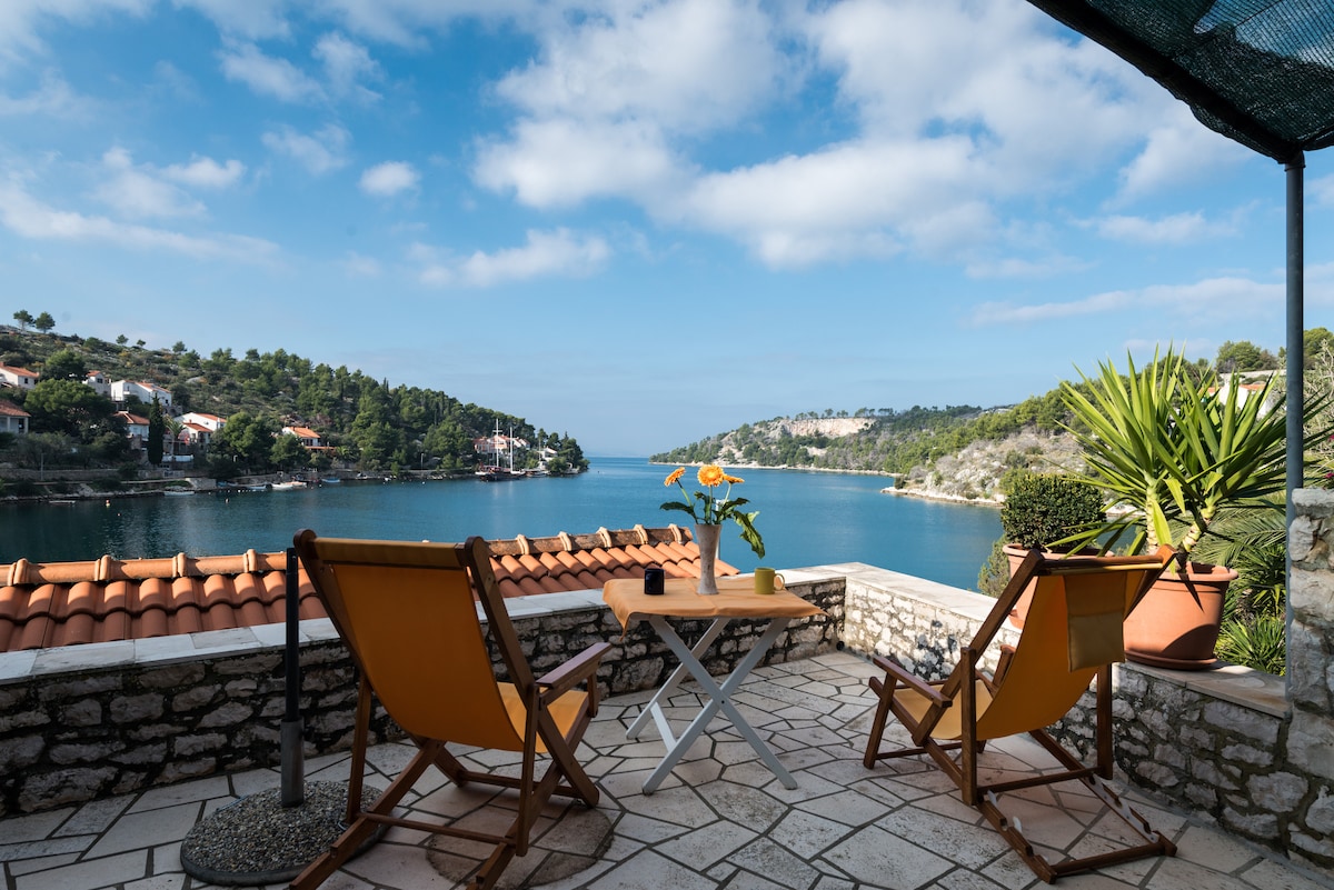 A terrace offers a water view framed by lush greenery and hills. Two orange chairs sit around a small white table, inviting relaxation. Sunlight illuminates the stone-paved surface, enhancing the outdoor space's appeal. The sky displays soft clouds against a bright backdrop.