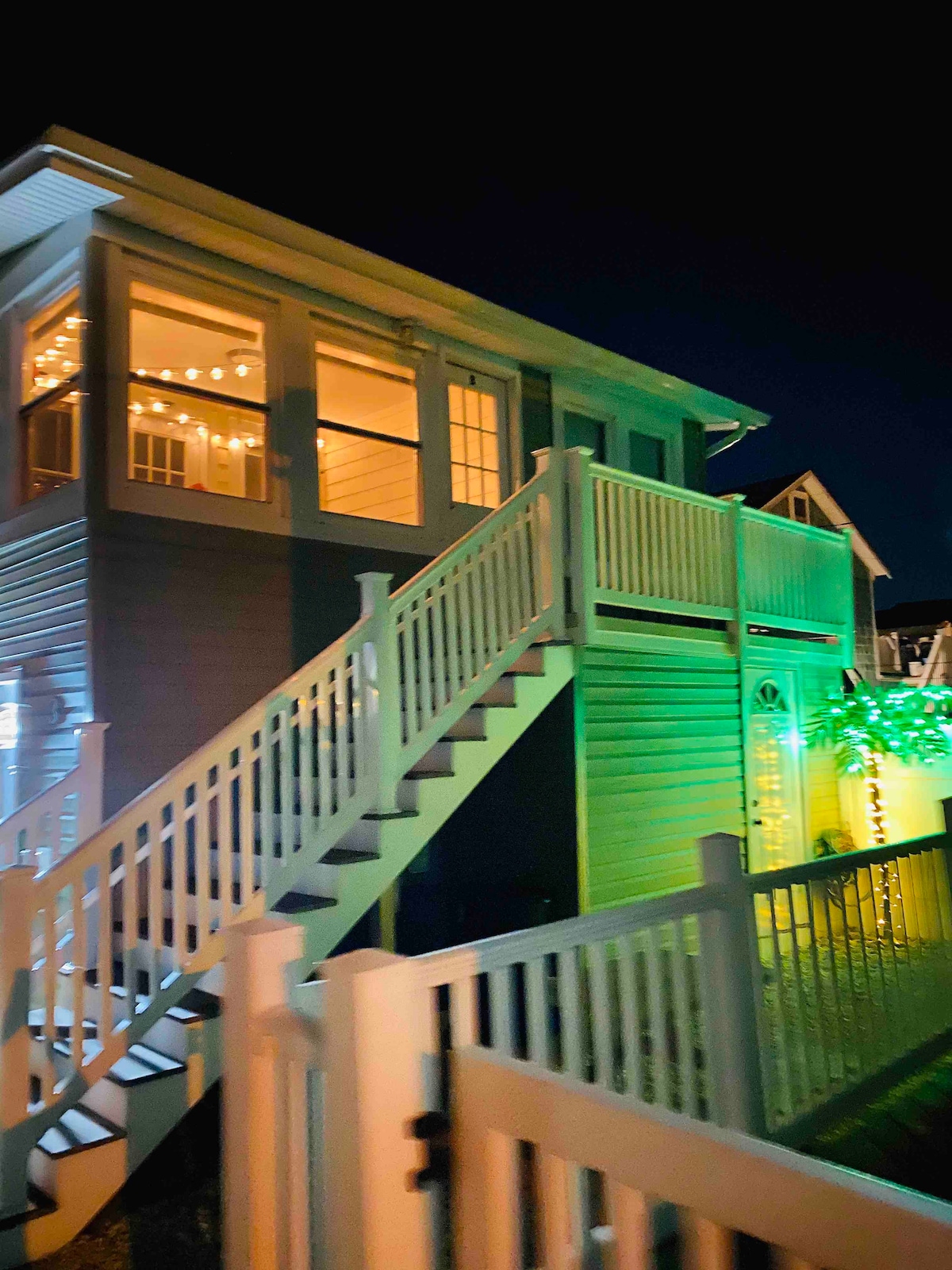 The exterior of the two-story house is illuminated at night, showcasing a staircase leading to the second floor. Decorative string lighting accentuates the enclosed porch's numerous windows, enhancing the inviting appearance of the property.