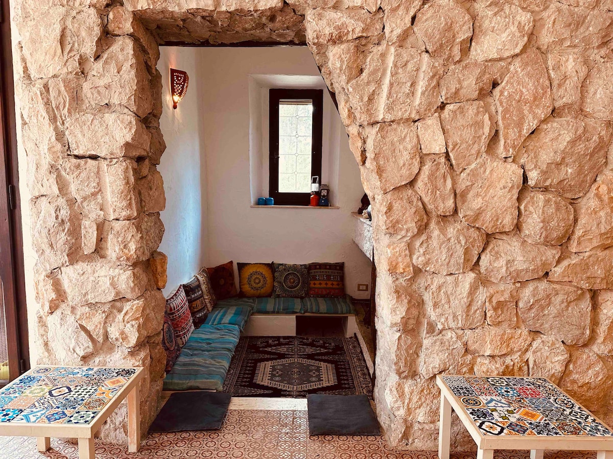 A cozy seating area is framed by a natural stone arch. Colorful cushions are arranged along a built-in bench, complemented by patterned rugs and small tables. A window provides natural light, enhancing the inviting atmosphere of the space.