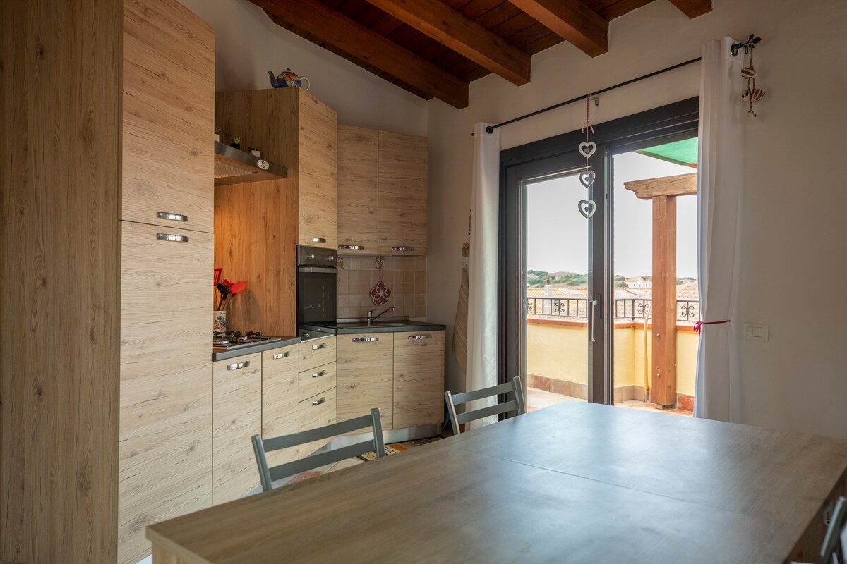 A modern kitchen area features light wood cabinetry and appliances, maintaining a clean and organized appearance. A wooden table with several chairs invites gatherings. Large sliding doors open to reveal a panoramic terrace, allowing natural light to brighten the space.