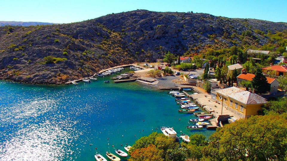 An expansive view of a serene bay features a pebbled beach with small boats anchored along the shore. Surrounding hills provide a natural backdrop, while colorful buildings and trees create a harmonious coastal scene under bright blue skies.