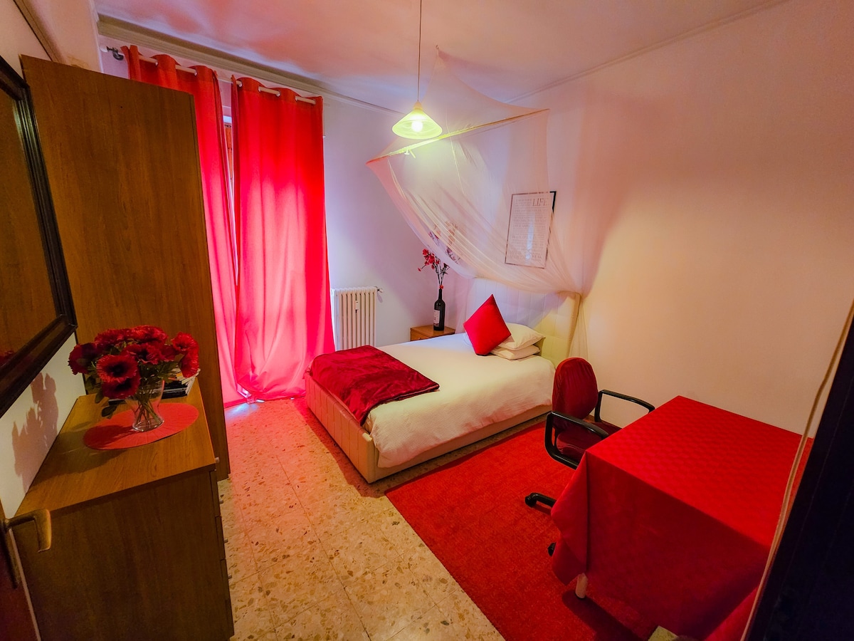 A bright and inviting room features a spacious bed draped with a canopy, complemented by red accents in curtains and decor. A small table and chair are positioned nearby, while a cabinet offers additional storage. Soft lighting enhances the overall warmth of the space.