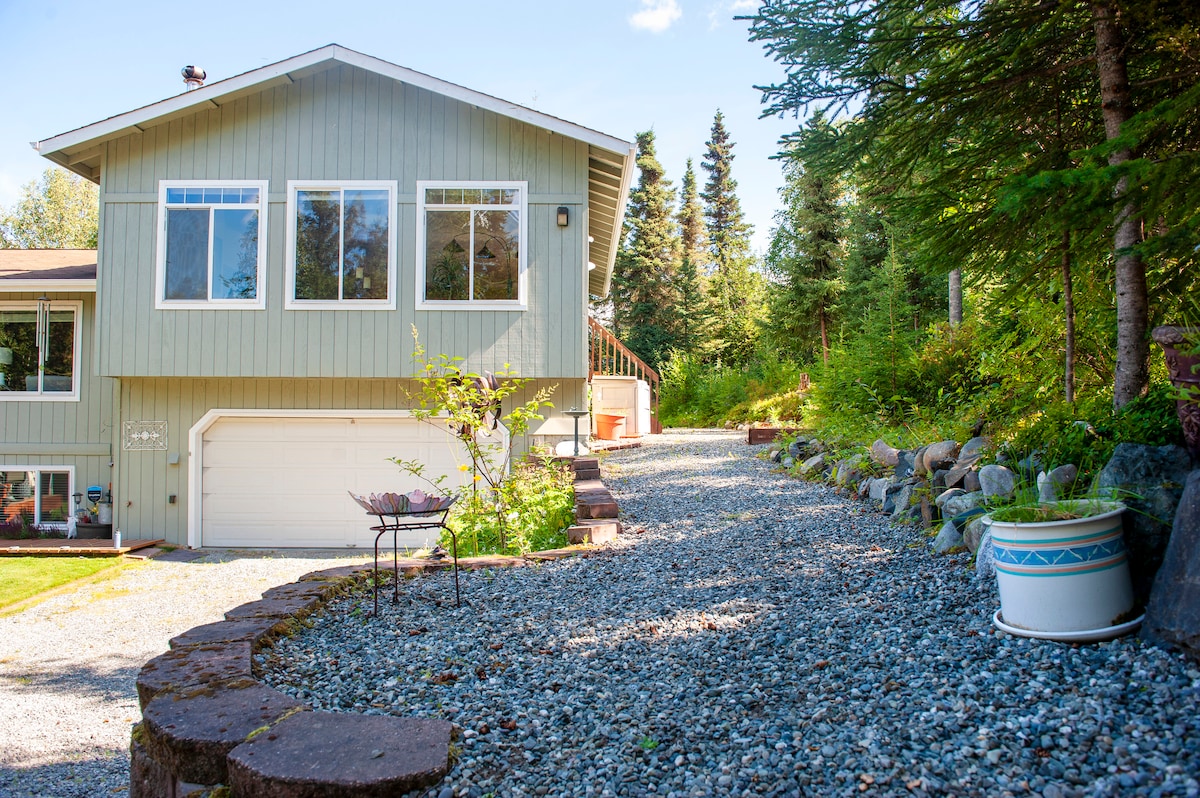 The exterior view of a two-story house is presented, featuring a gravel driveway leading to the entrance. Large windows enhance the facade, allowing natural light to enter. Surrounding trees and landscaping provide a sense of privacy in the quiet neighborhood.