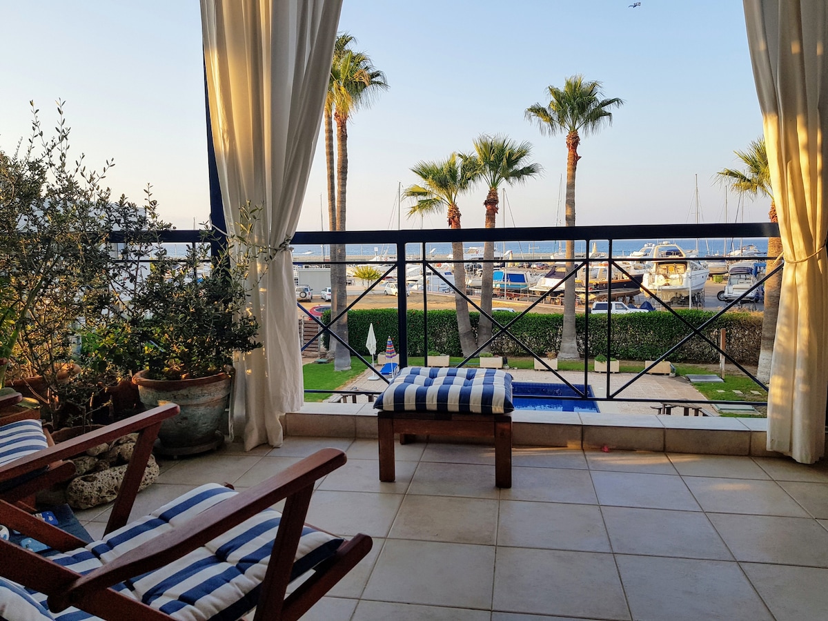 A large balcony is presented with a view of Latsi Marina. Palm trees frame the scene, and boats are visible in the water. There are two wooden lounge chairs with striped cushions facing the view, enhancing a sense of relaxation.