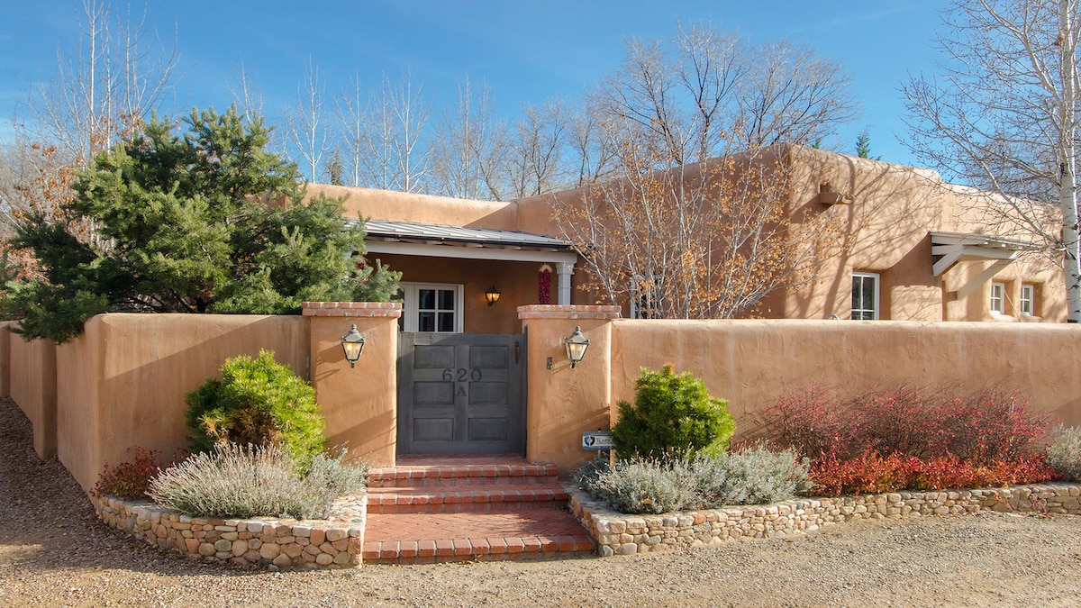 A charming adobe exterior is framed by lush greenery and colorful plants. The entrance features a rustic wooden door with a welcome sign, flanked by two wall sconces. A cobblestone pathway leads up to the door, creating a warm and inviting approach.