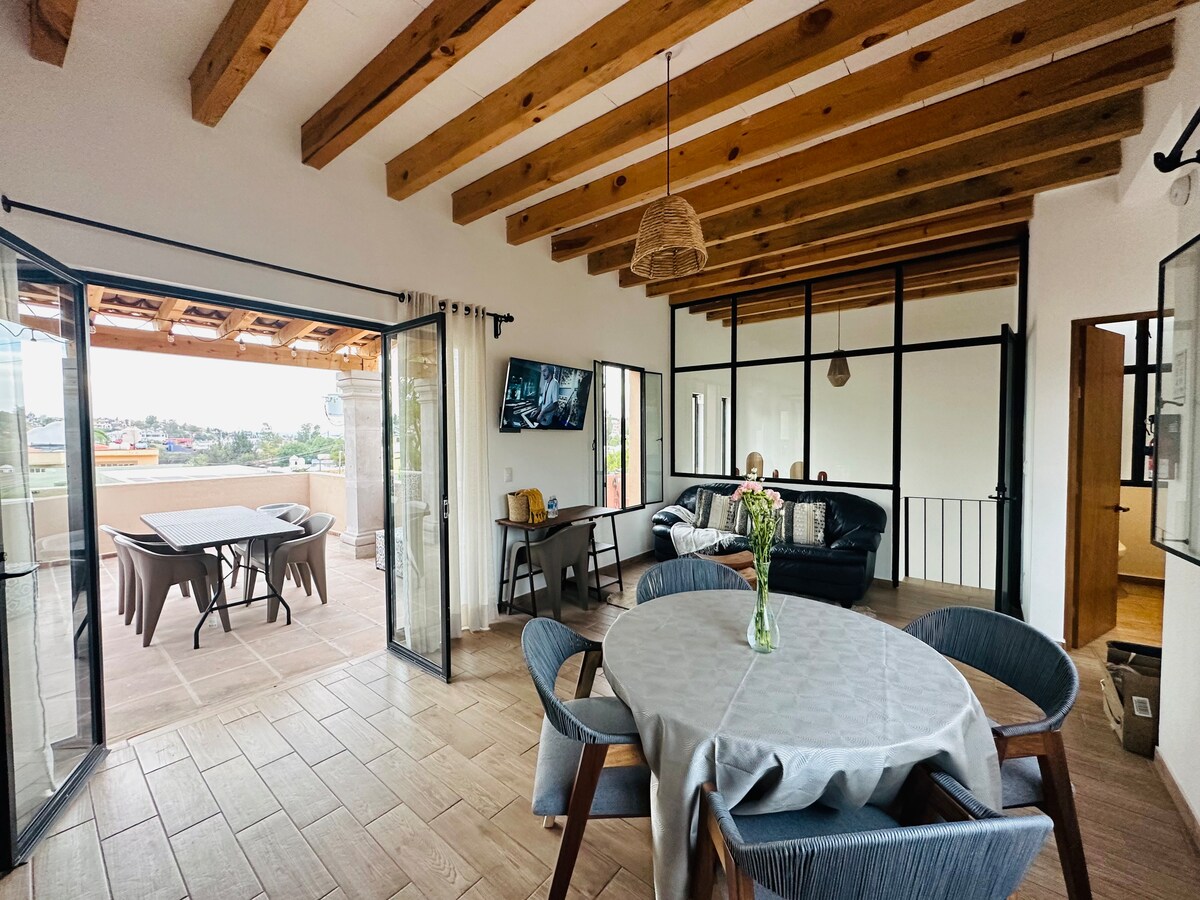 The open living area features wood beam ceilings, a circular dining table set for four, and a comfortable black sofa. A television is mounted on the wall opposite a balcony door, leading to an outdoor seating area with a table and chairs.