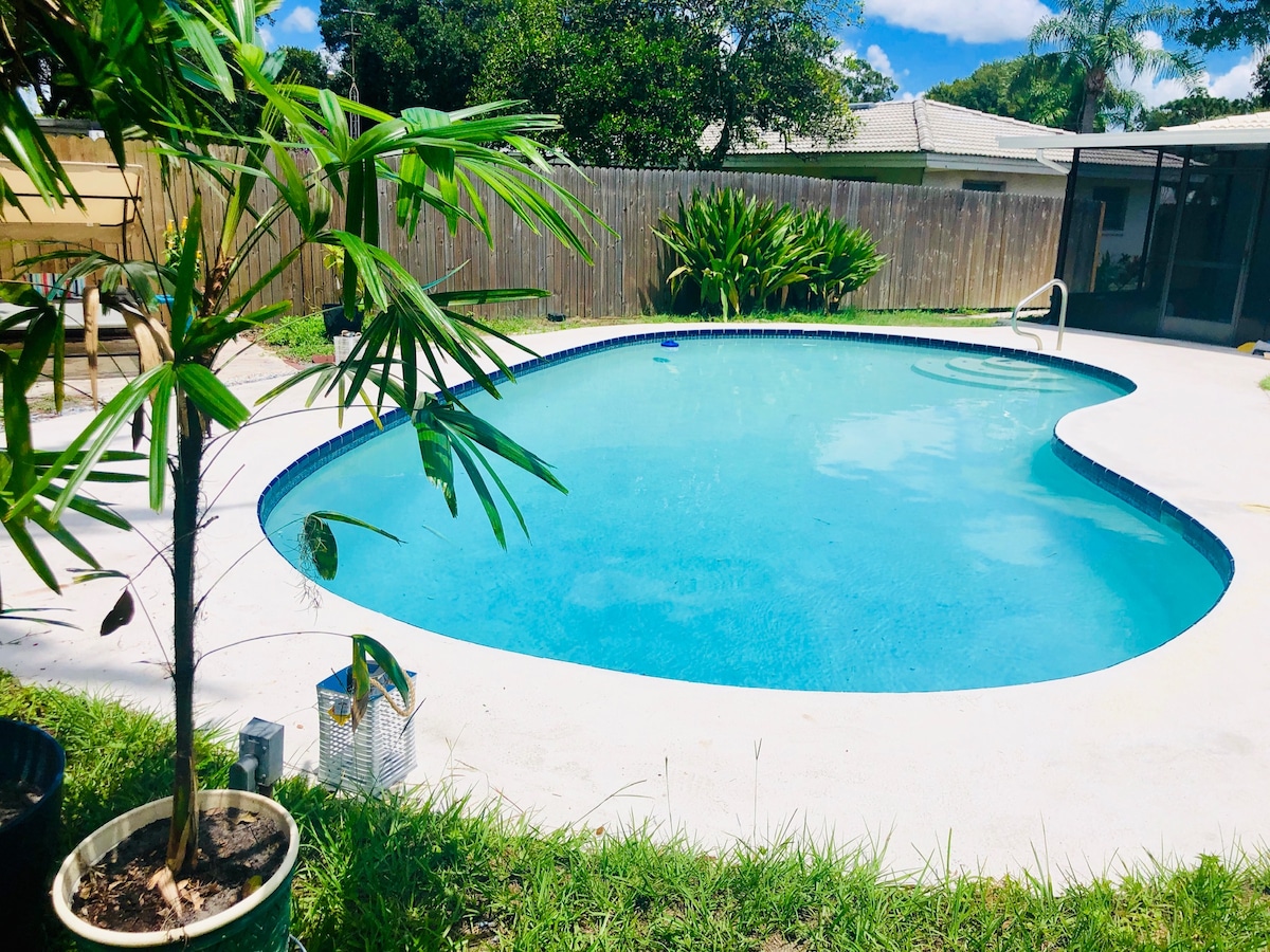 A clear swimming pool is set in a well-kept outdoor area, surrounded by lush greenery. The pool is framed by a light-colored deck, enhancing its inviting appearance. A small palm tree adds a touch of tropical ambiance, offering a peaceful retreat.